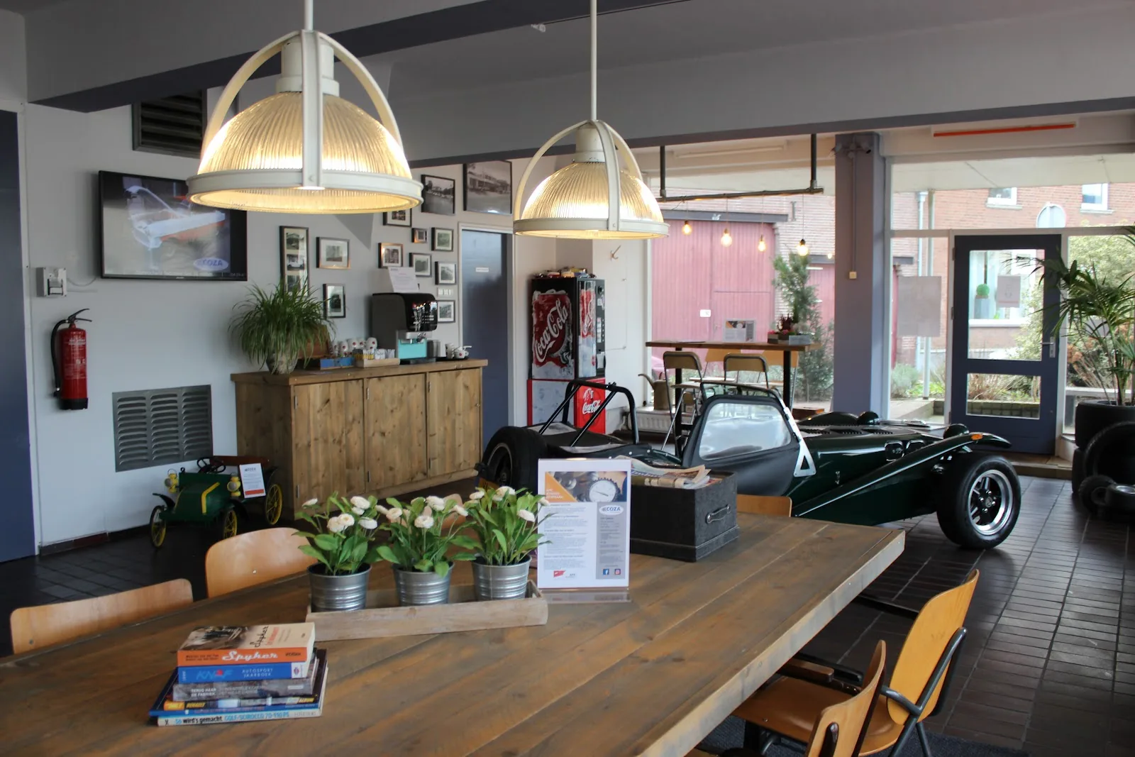 De wachtruimte van COZA Autokeuringen met een lange houten tafel, koffiehoek en een klassieke groene sportwagen